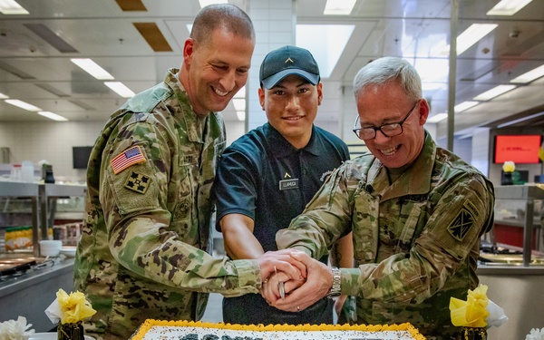 Three individuals — two in U.S. Army combat uniforms and one in civilian attire wearing a cap with a rank insignia — are jointly cutting a large sheet cake decorated with 'Army Birthday' text and gold frosting. The event appears to be an Army Birthday celebration held in a military dining facility (DFAC). Black and gold balloons and decorative elements reinforce the celebratory theme.