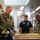 247th Army birthday cake cutting ceremony Three individuals are gathered around a large decorated sheet cake in what appears to be a military dining facility. A U.S. Army soldier in camouflage uniform is cutting the cake while two others look on, suggesting a celebratory military event. Yellow and black balloons are visible in the background, indicating a formal celebration.