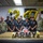 247th Army birthday cake cutting ceremony A group of U.S. Army personnel pose for a celebratory photo marking the Army's 247th birthday. The group includes two soldiers in Army Combat Uniforms (ACUs) flanking a team of culinary staff wearing black uniforms with aprons. A decorated cake and gold 'ARMY' lettering are displayed on a table in the foreground.