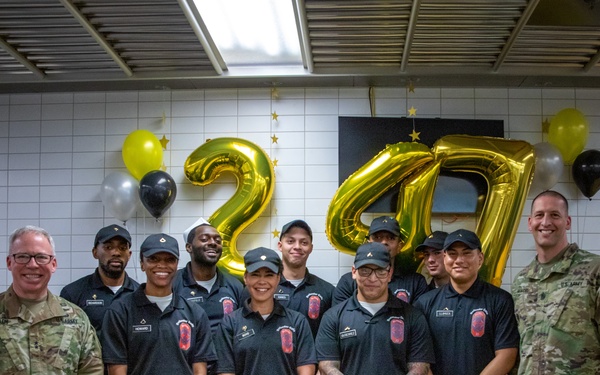 A group of U.S. Army personnel pose for a celebratory photo marking the Army's 247th birthday. The group includes two soldiers in Army Combat Uniforms (ACUs) flanking a team of culinary staff wearing black uniforms with aprons. A decorated cake and gold 'ARMY' lettering are displayed on a table in the foreground.