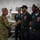 247th Army birthday cake cutting ceremony Two U.S. Army soldiers in MultiCam (OCP) uniforms are greeting staff members wearing dark navy uniforms bearing 'Defender Cafe' patches in what appears to be a military dining facility kitchen. A handshake is taking place between one of the soldiers and a cafe worker, suggesting an official visit or recognition event. A third cafe staff member is visible in the background observing the interaction.