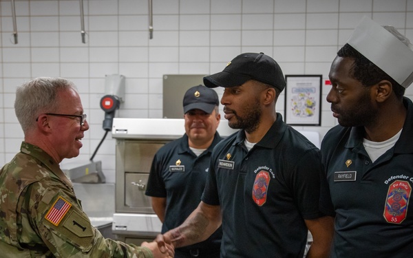 Two U.S. Army soldiers in MultiCam (OCP) uniforms are greeting staff members wearing dark navy uniforms bearing 'Defender Cafe' patches in what appears to be a military dining facility kitchen. A handshake is taking place between one of the soldiers and a cafe worker, suggesting an official visit or recognition event. A third cafe staff member is visible in the background observing the interaction.