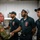 247th Army birthday cake cutting ceremony A U.S. Army officer in camouflage uniform is shaking hands with personnel wearing black 'Defender Cafe' polo shirts, likely in a military dining facility kitchen. The interaction appears to be a formal greeting or recognition visit. Three staff members wearing matching uniforms with name tags are visible, standing in a line.