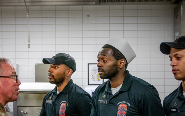 A U.S. Army officer in camouflage uniform is shaking hands with personnel wearing black 'Defender Cafe' polo shirts, likely in a military dining facility kitchen. The interaction appears to be a formal greeting or recognition visit. Three staff members wearing matching uniforms with name tags are visible, standing in a line.