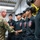 247th Army birthday cake cutting ceremony A U.S. Army officer in multicam/OCP uniform is shaking hands with a group of individuals wearing matching black polo shirts bearing an emblem and the number '35', likely fire department or emergency services personnel. The setting appears to be an indoor facility, possibly a kitchen or service area, decorated with balloons in the background. The interaction appears ceremonial or congratulatory in nature.