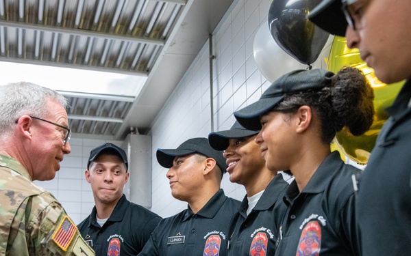 A U.S. Army officer in multicam/OCP uniform is shaking hands with a group of individuals wearing matching black polo shirts bearing an emblem and the number '35', likely fire department or emergency services personnel. The setting appears to be an indoor facility, possibly a kitchen or service area, decorated with balloons in the background. The interaction appears ceremonial or congratulatory in nature.
