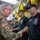 247th Army birthday cake cutting ceremony A U.S. Army soldier in OCP (Operational Camouflage Pattern) uniform is shaking hands with a person wearing a black 'Defender Gate Fire Police' uniform bearing the name 'Garmendia' and number 33. The setting appears to be a formal recognition or graduation ceremony, decorated with gold and black balloons and star decorations. Several other individuals in matching black uniforms are lined up in the background.
