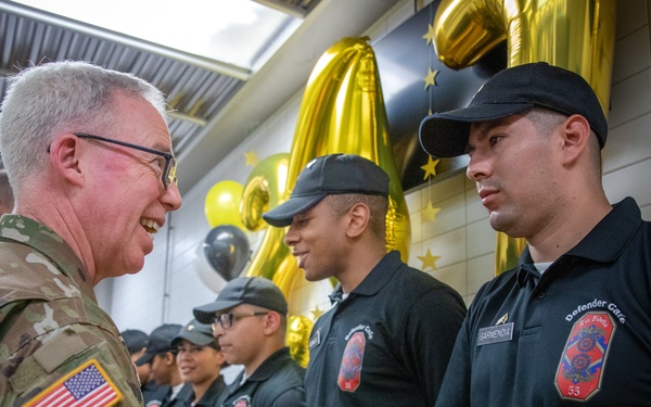 A U.S. Army soldier in OCP (Operational Camouflage Pattern) uniform is shaking hands with a person wearing a black 'Defender Gate Fire Police' uniform bearing the name 'Garmendia' and number 33. The setting appears to be a formal recognition or graduation ceremony, decorated with gold and black balloons and star decorations. Several other individuals in matching black uniforms are lined up in the background.
