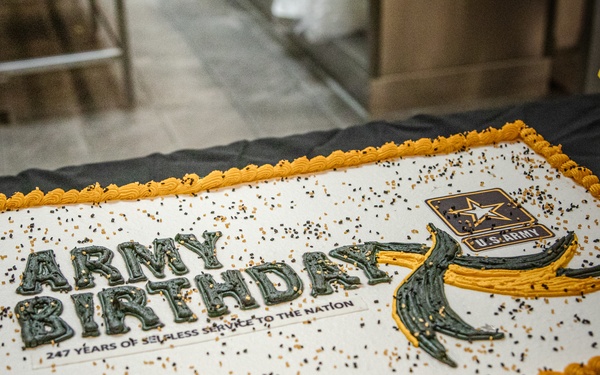 A large rectangular sheet cake celebrating the U.S. Army's 247th Birthday is displayed on a black tablecloth. The cake features gold and dark green frosting decorations, the official U.S. Army star logo, and the text '247 Years of Selfless Service to the Nation.' The setting appears to be an indoor military celebration event.