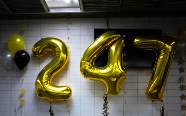 The image depicts a celebratory display for the U.S. Army's 247th birthday, featuring large gold foil number balloons spelling '247' against a tiled wall. A decorated cake with '247' candles sits on a table in front of large black letters spelling 'ARMY', surrounded by gold glitter tablecloth and floral arrangements. The color scheme is consistent with Army branding: black, gold, and white.