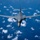 A gray military strategic bomber aircraft is photographed from above and slightly ahead, flying at high altitude over a deep blue ocean with scattered white cumulus clouds below. The aircraft displays a variable-sweep wing configuration with wings in the extended position. The image is a professional aerial photograph likely taken from a companion aircraft.