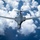 A U.S. Air Force B-1B Lancer supersonic strategic bomber is photographed from below and slightly ahead, flying at altitude against a backdrop of cumulus clouds and blue sky. The aircraft is shown in a swept-wing configuration with wings extended, displaying its characteristic variable-geometry design. The image appears to be an official or professional aerial photograph taken from another aircraft.