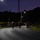 Headquarters Battalion Conditioning Hike A nighttime military formation or march is taking place on a paved road, illuminated by street lights. A group of uniformed personnel are moving along the road, with one individual visibly carrying a red and yellow flag or guidon. The scene appears to be on a military installation, with a high-voltage transmission tower and forested hills visible in the background.
