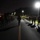 Headquarters Battalion Conditioning Hike A large column of military personnel conducting a nighttime road march along a paved road illuminated by street lights. Soldiers are wearing combat gear, rucksacks, and reflective safety vests, marching in formation away from the camera. A red flag or guidon is visible near the front of the formation.