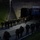 Headquarters Battalion Conditioning Hike A large group of military personnel in camouflage uniforms and carrying heavy rucksacks are marching through and emerging from an underpass or tunnel at night. The scene is illuminated by a single overhead street lamp, casting dramatic shadows on the concrete structure. A clearance sign on the tunnel reads 'MAX CLEARANCE 13 FT / 4.5 M'.