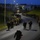 Headquarters Battalion Conditioning Hike A large column of military personnel conducting a night march or ruck march along a paved road on what appears to be a military installation. Soldiers are carrying full backpacks/rucksacks and are organized in formation along both sides of the road. The scene is illuminated by street lights against a deep blue twilight sky.