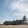 A U.S. Air Force C-130 Hercules transport aircraft belonging to the Ohio Air National Guard is parked on a tarmac with its propellers spinning. Military personnel are visible on both sides of the aircraft, suggesting pre-flight or post-flight ground operations. The image was taken on a clear, sunny day with scattered cirrus clouds and contrails visible in the sky.