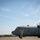 A U.S. Air Force C-130 Hercules transport aircraft assigned to the Ohio Air National Guard is being marshaled on a flight line tarmac. A uniformed airman in camouflage uniform is directing the aircraft using marshaling wands with both arms raised. An air traffic control tower and an American flag are visible in the background.