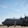 A U.S. Air Force C-130 Hercules transport aircraft assigned to the Ohio Air Guard is shown on a tarmac with engines running, as indicated by the spinning propellers. A uniformed service member stands in front of the aircraft in a saluting position, likely performing marshaling or ceremonial duties. An air traffic control tower is visible in the background to the left.