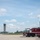 The image shows an airport or military airfield with emergency fire response vehicles parked near a hangar. A control tower is visible in the background, and an aircraft appears to be on approach or takeoff in the far left. The scene suggests routine airfield operations with fire/rescue standby.
