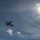 A low-angle photograph captures a multi-engine turboprop aircraft in flight against a blue sky with scattered clouds. The sun is prominently visible in the upper right corner, creating a strong lens flare effect. The aircraft appears to be a large military or cargo-type turboprop, photographed from directly below.