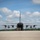 A large military transport aircraft, consistent with a C-130 Hercules, is photographed from a rear perspective on a concrete tarmac with its rear cargo ramp open. Several personnel in flight suits are walking out from the open cargo bay. The image is taken on a partly cloudy day with green tree lines visible in the background.