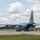A U.S. Air National Guard C-130 Hercules transport aircraft is parked on a tarmac with its rear cargo ramp lowered. A group of approximately 6-8 military personnel in flight suits and combat uniforms are walking away from the aircraft's rear ramp. The setting appears to be a military airfield surrounded by trees under partly cloudy skies.