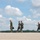 A group of six military personnel in flight suits and combat uniforms are walking across what appears to be an airfield tarmac. Most are wearing olive drab flight suits, with one individual in a camouflage (MultiCam/OCP) uniform. They appear to be moving purposefully, possibly after completing or preparing for a flight mission.