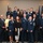 A formal group photograph featuring approximately 20 individuals, the majority of whom are wearing United States Air Force dress uniforms. The group appears to be gathered for an official ceremony or recognition event, posed in two rows in front of American and Air Force flags. Several civilians in business casual attire are interspersed among the uniformed personnel.