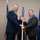 A formal military ceremony showing a U.S. Air Force officer in dress blues passing or receiving a guidon (unit flag on a staff) to/from a civilian in a dark suit. A third uniformed individual stands in the background. The setting appears to be an indoor ceremonial venue with U.S. and Air Force flags visible.