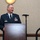 A U.S. Air Force officer in dress blue uniform stands at a wooden podium, appearing to deliver a speech or remarks at a formal military event. His name tag reads 'McClain' and he wears multiple service ribbons and badges on his uniform. A microphone on a stand is positioned nearby.