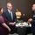 Three people are engaged in conversation at what appears to be a formal dining event or military luncheon. A uniformed military officer in US Air Force dress blues is speaking with a man in a business suit and a woman in a purple lace dress. A fourth person in a yellow polo shirt is visible in the background observing the scene.