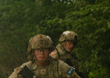 Two U.S. Army soldiers in full combat gear are conducting what appears to be a field training exercise on a dirt path surrounded by dense vegetation. The foreground soldier is in a kneeling tactical position holding an M4-style carbine rifle, while a second soldier stands behind in a similar posture. Both wear Operational Camouflage Pattern (OCP) uniforms with helmets and protective eyewear.