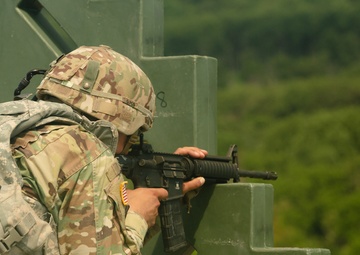 A U.S. Army soldier in Operational Camouflage Pattern (OCP) uniform is firing an M4/M16-style carbine from behind a green concrete barrier at what appears to be a military training range. A spent brass casing is visibly ejecting from the weapon, captured mid-flight to the upper right. The lush green treeline in the background and outdoor setting suggest a live-fire training exercise.