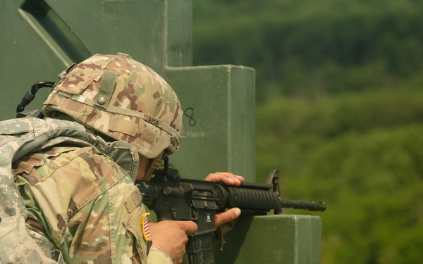 A U.S. Army soldier in Operational Camouflage Pattern (OCP) uniform is firing an M4/M16-style carbine from behind a green concrete barrier at what appears to be a military training range. A spent brass casing is visibly ejecting from the weapon, captured mid-flight to the upper right. The lush green treeline in the background and outdoor setting suggest a live-fire training exercise.