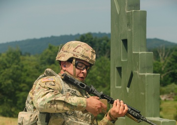 A U.S. Army soldier in full combat gear is kneeling behind a green concrete barrier structure on a gravel training range. The soldier is holding a rifle in a low-ready or transitioning position, appearing to be engaged in a tactical shooting exercise. The setting is an outdoor military training facility with wooded hills visible in the background.