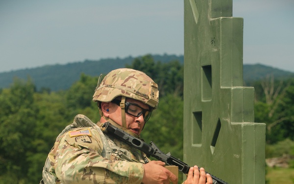 A U.S. Army soldier in full combat gear is kneeling behind a green concrete barrier structure on a gravel training range. The soldier is holding a rifle in a low-ready or transitioning position, appearing to be engaged in a tactical shooting exercise. The setting is an outdoor military training facility with wooded hills visible in the background.