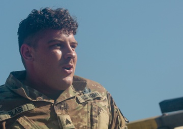A U.S. Army soldier wearing Operational Camouflage Pattern (OCP) uniform is photographed from a low angle against a clear blue sky. The soldier, identified by the name tape 'BLAIR' on the left chest, holds the rank of Specialist (SPC) as indicated by the rank insignia on the collar. The soldier appears to be resting or observing something off-camera near a wooden structure.