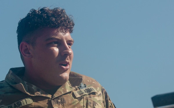 A U.S. Army soldier wearing Operational Camouflage Pattern (OCP) uniform is photographed from a low angle against a clear blue sky. The soldier, identified by the name tape 'BLAIR' on the left chest, holds the rank of Specialist (SPC) as indicated by the rank insignia on the collar. The soldier appears to be resting or observing something off-camera near a wooden structure.
