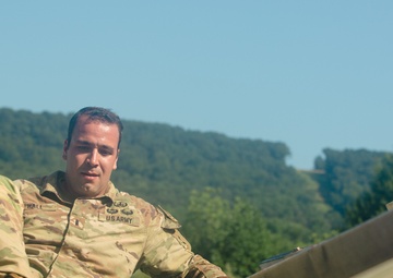 A U.S. Army soldier in OCP (Operational Camouflage Pattern) uniform is navigating what appears to be an obstacle course, climbing over a wooden barrier or wall. The soldier's name tape reads 'HALL' and the chest tape reads 'U.S. ARMY'. The setting is an outdoor training environment with wooded hills and clear blue sky in the background.