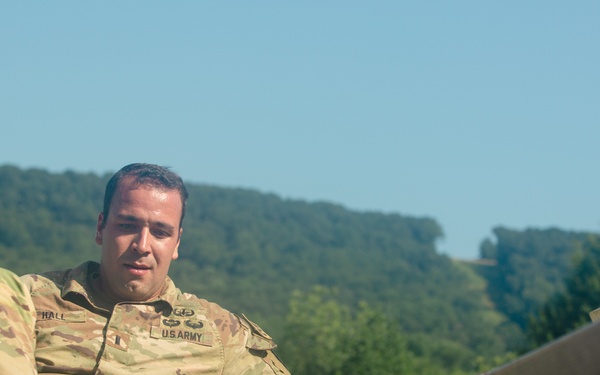 A U.S. Army soldier in OCP (Operational Camouflage Pattern) uniform is navigating what appears to be an obstacle course, climbing over a wooden barrier or wall. The soldier's name tape reads 'HALL' and the chest tape reads 'U.S. ARMY'. The setting is an outdoor training environment with wooded hills and clear blue sky in the background.