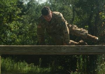 A U.S. Army soldier in full camouflage uniform is navigating a military obstacle course, specifically crawling or vaulting over a horizontal wooden beam barrier. The soldier is wearing patches including an American flag and what appears to be an 'EOD' (Explosive Ordnance Disposal) tab. The setting is an outdoor training facility with a gravel ground surface and wooded background.