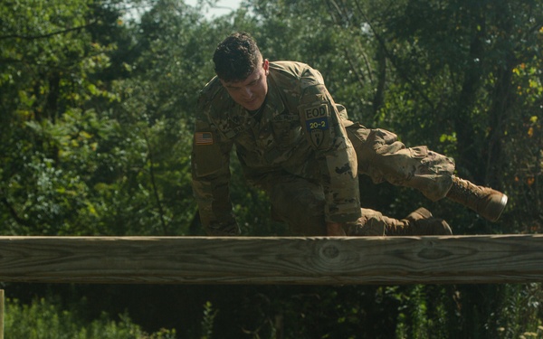 A U.S. Army soldier in full camouflage uniform is navigating a military obstacle course, specifically crawling or vaulting over a horizontal wooden beam barrier. The soldier is wearing patches including an American flag and what appears to be an 'EOD' (Explosive Ordnance Disposal) tab. The setting is an outdoor training facility with a gravel ground surface and wooded background.