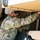 A U.S. Army soldier in OCP (Operational Camouflage Pattern) uniform is inspecting or performing maintenance on the undercarriage of a military vehicle. The soldier is wearing sunglasses and reaching under the tan/desert-colored vehicle, examining components including chains and mechanical parts. The image appears to be taken during a maintenance or pre-operation check.