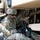 Two U.S. Army soldiers in OCP (Operational Camouflage Pattern) uniforms are working underneath or alongside a large military vehicle, handling chains and inspecting mechanical components. The soldiers appear to be performing maintenance, rigging, or recovery operations on the tan-colored military vehicle. The image captures close-up detail of the undercarriage area including axles, drive shafts, and large off-road tires.