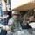 Two U.S. Army soldiers in full combat uniforms and helmets are working underneath or alongside a large military vehicle, handling recovery or rigging hardware including a shackle and chain. The soldier on the right has a name tape reading 'ORANGE' and both wear OCP (Operational Camouflage Pattern) uniforms with U.S. flag patches. The scene appears to depict a vehicle recovery or towing operation.