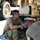 Two military personnel in olive drab/OCP uniforms are performing maintenance or inspection on the undercarriage of a tan/desert-colored military vehicle. One soldier is crouching and observing while the other is leaning underneath the vehicle to inspect or work on a component. The vehicle appears to be a large military truck with a tow hook or coupling mechanism visible.