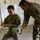 Two U.S. Army soldiers in OCP (Operational Camouflage Pattern) uniforms are engaged in what appears to be a medical training exercise indoors. The kneeling soldier, identified by name tape as 'BARRETT,' is handling what appears to be a bandage or medical wrap, while a second soldier leans over to provide instruction or guidance. The setting appears to be a training room with whiteboard briefing charts visible in the background.