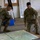 Two U.S. Army soldiers in OCP (Operational Camouflage Pattern) uniforms are kneeling on a concrete floor examining a large topographic map. The setting appears to be a military operations or planning room, with whiteboards covered in handwritten notes and lists visible in the background. This appears to be a map reconnaissance or mission planning activity.