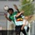 Two female soccer players are competing for a header during a match between Cameroon and Belgium. The player in the green Cameroon kit is winning the aerial duel, making contact with the ball using her head, while the Belgian player (number 16) in a light blue and black kit is closely contesting the challenge. The action is captured mid-air with dynamic motion blur on the Belgian player's ponytail.